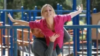 Mulher loira sorridente é erguida por homem barbudo em playground, braços abertos, simulando voo. Clima alegre.