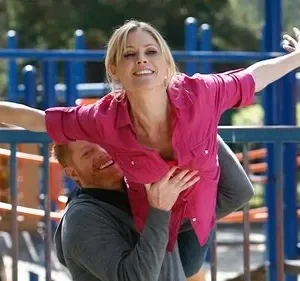 Mulher loira sorridente é erguida por homem barbudo em playground, braços abertos, simulando voo. Clima alegre.