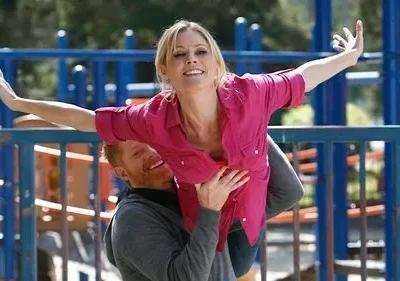 Mulher loira sorridente é erguida por homem barbudo em playground, braços abertos, simulando voo. Clima alegre.