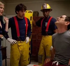 Mulher loira fala com dois paramédicos. Um homem sentado faz careta de dor. Cena de emergência cômica em casa.