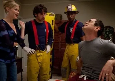 Mulher loira fala com dois paramédicos. Um homem sentado faz careta de dor. Cena de emergência cômica em casa.