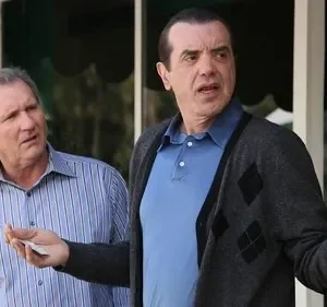 Dois homens de meia-idade conversam ao ar livre. Um de camisa listrada parece confuso, o outro de casaco gesticula expressivamente.