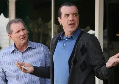 Dois homens de meia-idade conversam ao ar livre. Um de camisa listrada parece confuso, o outro de casaco gesticula expressivamente.