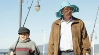 Homem de chapéu azul e menino de gorro seguram varas de pescar num cais, sob céu claro. Ambos têm expressões sérias.