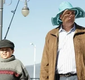 Homem de chapéu azul e menino de gorro seguram varas de pescar num cais, sob céu claro. Ambos têm expressões sérias.