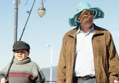 Homem de chapéu azul e menino de gorro seguram varas de pescar num cais, sob céu claro. Ambos têm expressões sérias.