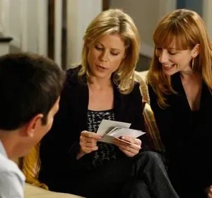 No sofá, uma mulher loira mostra fotos a uma ruiva sorridente. Um homem de costas observa a interação. Ambiente doméstico leve e amigável.