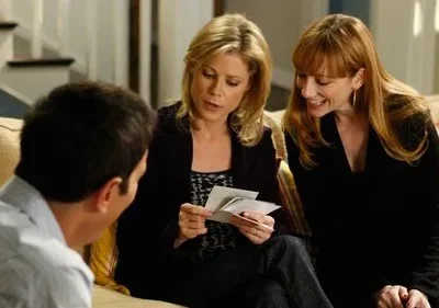 No sofá, uma mulher loira mostra fotos a uma ruiva sorridente. Um homem de costas observa a interação. Ambiente doméstico leve e amigável.