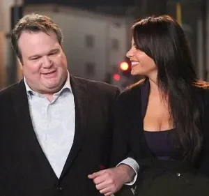 Um homem sorridente e uma mulher de cabelos escuros, de braços dados, olham um para o outro em uma rua noturna iluminada.