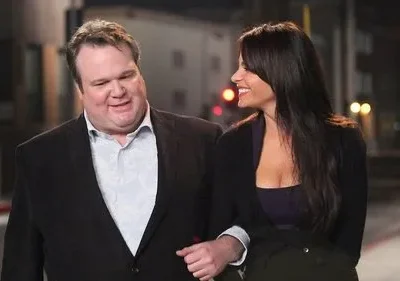 Um homem sorridente e uma mulher de cabelos escuros, de braços dados, olham um para o outro em uma rua noturna iluminada.