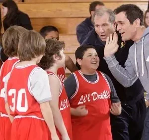 Um coach sorridente gesticula para meninos em uniformes de basquete vermelhos em quadra. Um menino ri abertamente. Clima divertido e esportivo.