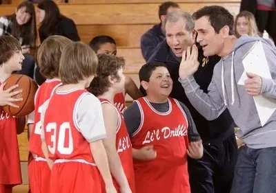 Um coach sorridente gesticula para meninos em uniformes de basquete vermelhos em quadra. Um menino ri abertamente. Clima divertido e esportivo.