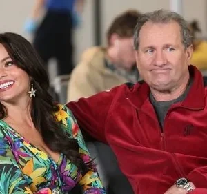 Uma mulher sorridente em vestido tropical e um homem com cara de poucos amigos em agasalho vermelho sentados em um aeroporto.