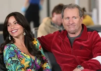 Uma mulher sorridente em vestido tropical e um homem com cara de poucos amigos em agasalho vermelho sentados em um aeroporto.