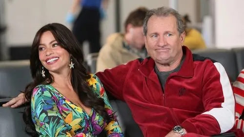 Uma mulher sorridente em vestido tropical e um homem com cara de poucos amigos em agasalho vermelho sentados em um aeroporto.