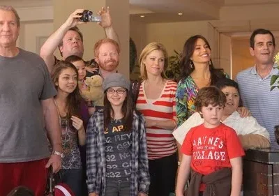 Grande família com adultos e crianças de várias idades sorri em um ambiente interno iluminado, transmitindo um clima feliz e de férias.