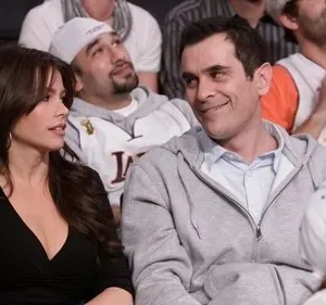 Mulher de cabelos escuros e homem sorridente em arquibancada. Ele a olha. Fãs atrás vestem camisas de basquete roxas e amarelas.