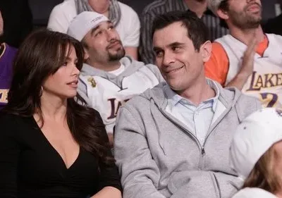 Mulher de cabelos escuros e homem sorridente em arquibancada. Ele a olha. Fãs atrás vestem camisas de basquete roxas e amarelas.