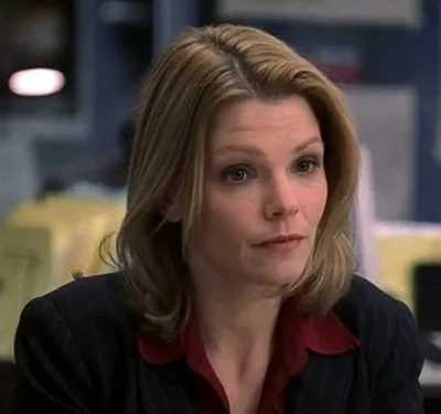 Mulher de cabelo loiro, blazer listrado escuro e camisa vermelha, com expressão séria, em um escritório.