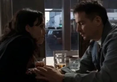 Um casal se olha intensamente em um bar, mãos dadas sobre a mesa com bebidas. O clima é íntimo e pensativo.