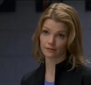 Uma mulher loira com expressão séria veste blazer escuro num ambiente interno com fundo cinza. Clima profissional.