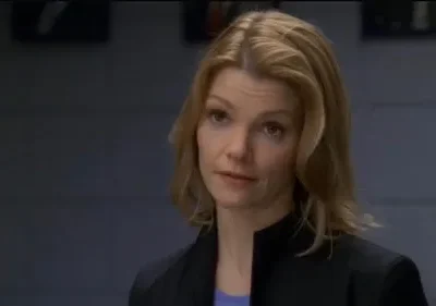 Uma mulher loira com expressão séria veste blazer escuro num ambiente interno com fundo cinza. Clima profissional.