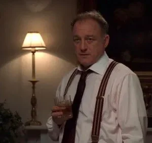 Homem de meia-idade, camisa, gravata e suspensórios, segura copo com bebida. Olhar sério em ambiente clássico pouco iluminado.