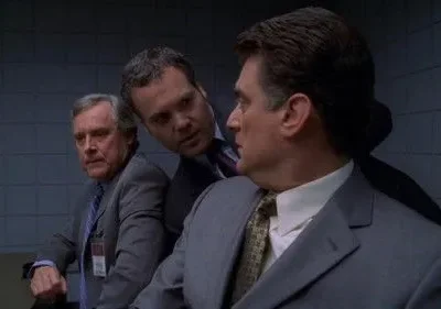 Três homens de terno em uma sala simples. Um se inclina para o outro, que parece sério. Clima tenso e formal.