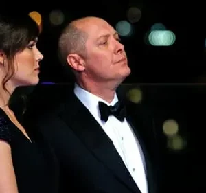 Um homem de smoking e uma mulher elegante de vestido preto olham em direções opostas à noite, com luzes de fundo desfocadas.