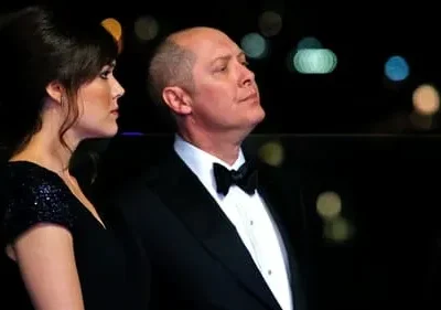 Um homem de smoking e uma mulher elegante de vestido preto olham em direções opostas à noite, com luzes de fundo desfocadas.