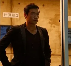 Homem asiático sério de blazer escuro em ambiente tecnológico. Monitores exibem código, criando um clima misterioso.