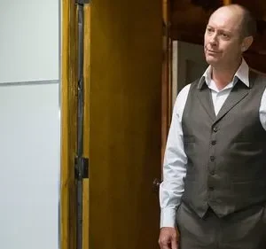 Um homem careca de colete escuro e camisa clara sorri levemente. Ele está em um interior com parede clara e detalhes de madeira.