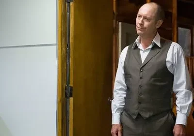 Um homem careca de colete escuro e camisa clara sorri levemente. Ele está em um interior com parede clara e detalhes de madeira.