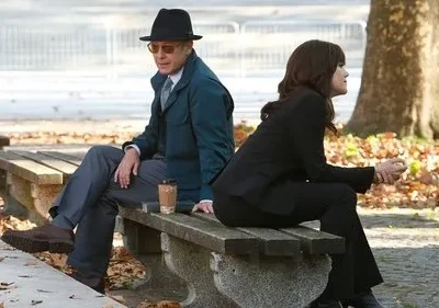 Um homem de chapéu e uma mulher em terno sentados num banco de parque com folhas de outono, olhando em direções opostas.
