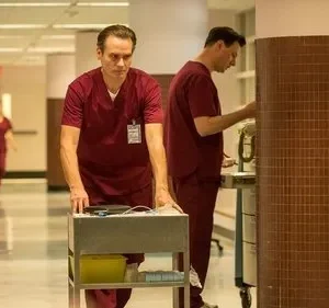 Homem em scrubs vermelhos empurra carrinho médico com seriedade em corredor hospitalar. Outros profissionais ao fundo.