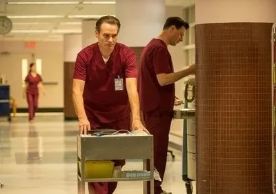 Homem em scrubs vermelhos empurra carrinho médico com seriedade em corredor hospitalar. Outros profissionais ao fundo.