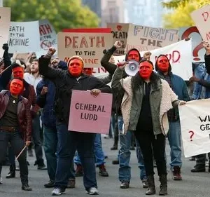 Grupo de manifestantes com rostos vermelhos marcha em uma rua. Seguram cartazes como Quem é General Ludd? e Morte à Máquina. Clima de revolta.