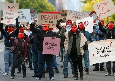 Grupo de manifestantes com rostos vermelhos marcha em uma rua. Seguram cartazes como Quem é General Ludd? e Morte à Máquina. Clima de revolta.