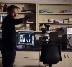 Homem barbudo de preto aponta arma num laboratório científico com equipamentos, incluindo microscópio e monitores. Cena de suspense.