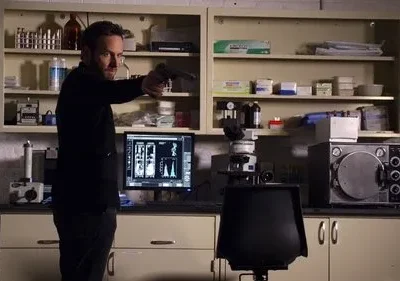 Homem barbudo de preto aponta arma num laboratório científico com equipamentos, incluindo microscópio e monitores. Cena de suspense.