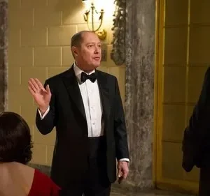 Um homem de smoking preto gesticula em um salão luxuoso, enquanto uma mulher de vestido vermelho e outro homem de smoking o observam de costas.