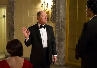 Um homem de smoking preto gesticula em um salão luxuoso, enquanto uma mulher de vestido vermelho e outro homem de smoking o observam de costas.