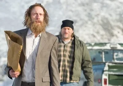 Dois homens em paisagem nevada. Um barbudo, de terno e saco de papel. O outro, de gorro e jaqueta xadrez. Ambiente frio e sério.