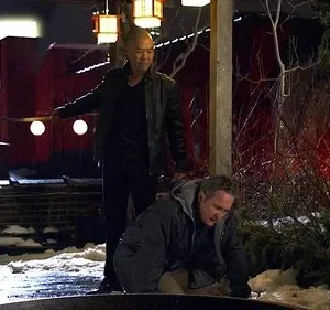 Um homem careca de jaqueta escura ameaça outro ajoelhado na neve, à noite. Fundo urbano com torre vermelha iluminada.