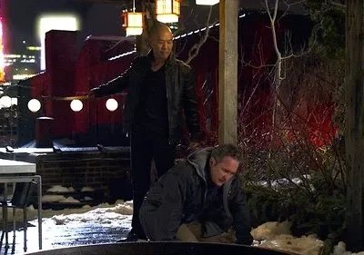 Um homem careca de jaqueta escura ameaça outro ajoelhado na neve, à noite. Fundo urbano com torre vermelha iluminada.