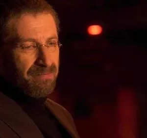 Homem barbudo de óculos e jaqueta, expressão séria, em ambiente escuro com luz vermelha ao fundo.