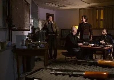 Homens sérios em sala escura, com rifles em primeiro plano e outras armas sobre a mesa. Ambiente tenso e clandestino.