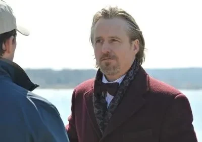 Um homem de casaco vinho e gravata borboleta olha pensativo, enquanto outro de boné está de costas, junto à água.