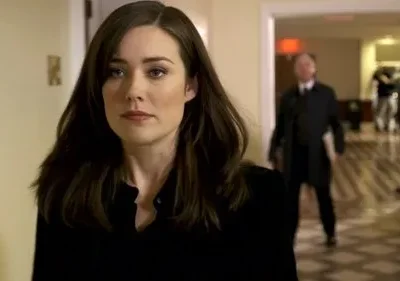 Jovem mulher de cabelo escuro, olhar sério, em corredor. No fundo, um homem de casaco e outras figuras borradas. Clima misterioso.