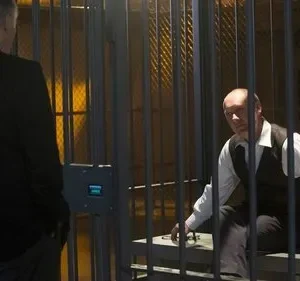 Um homem de costas de terno observa um homem careca com colete dentro de uma cela com barras. Clima tenso e sério.